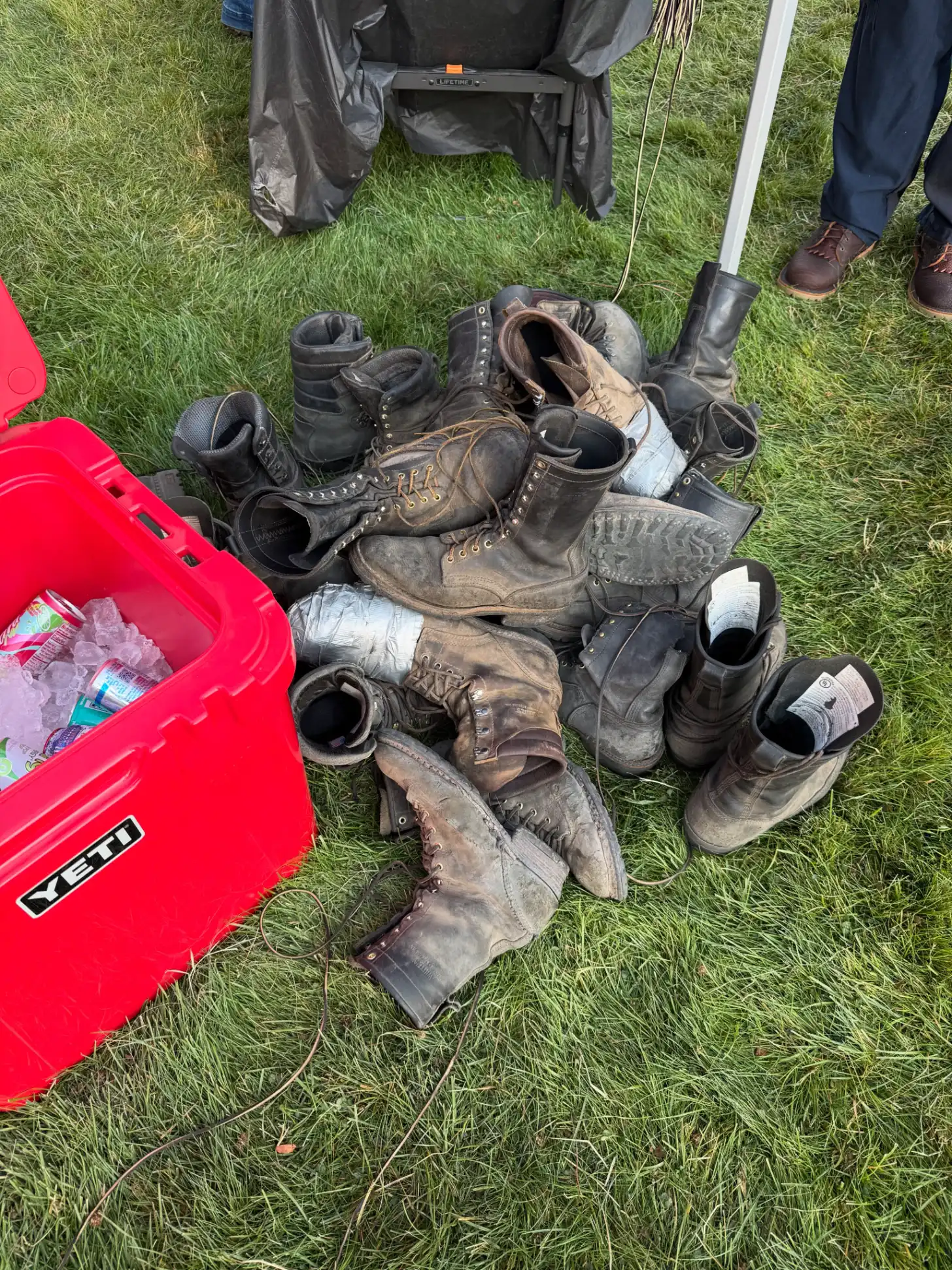 A collection of worn work boots and hiking boots arranged on grass next to a red cooler with people in work clothes standing nearby.
