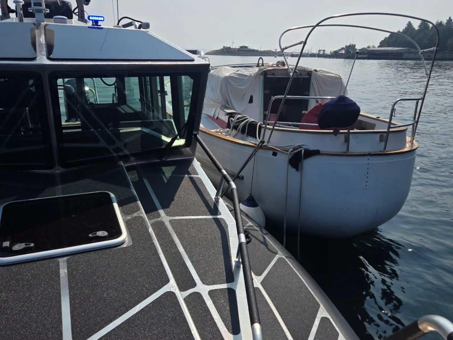 Two boats docked side by side on calm water. The boat on the left features a black cabin with windows, while the boat on the right is white with a protective cover and metal railings. Forested shoreline and industrial structures are visible in the background.