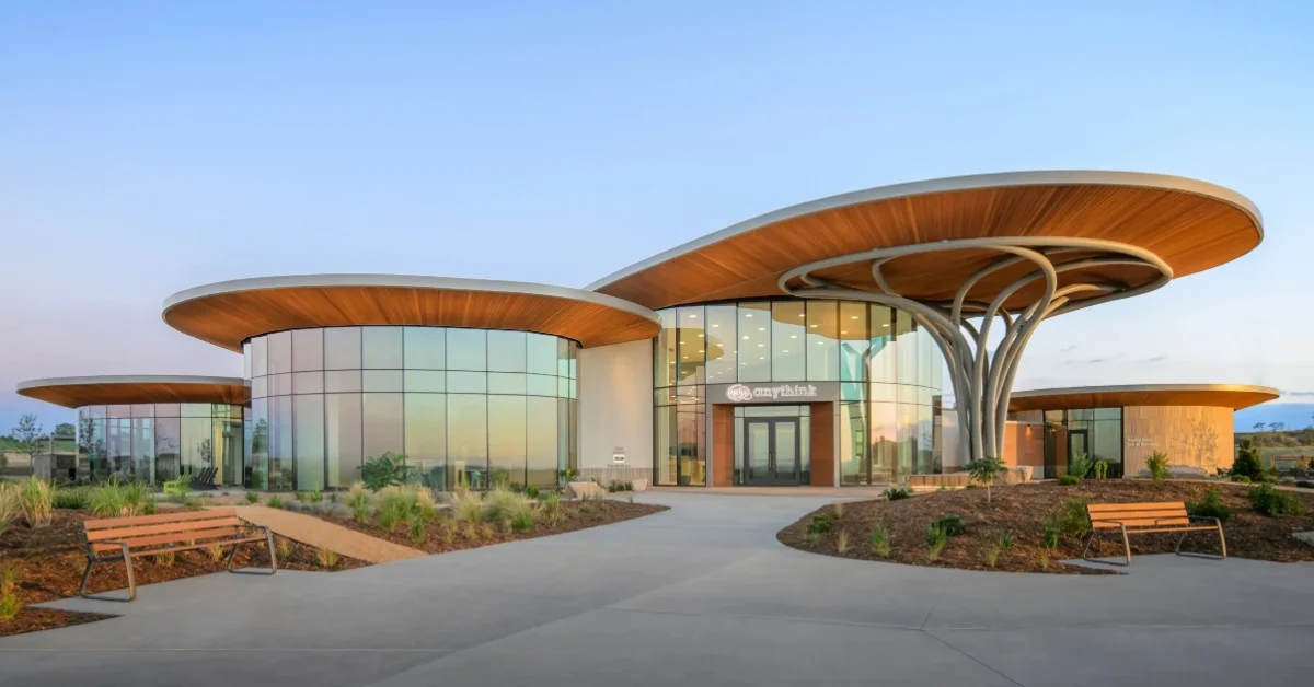 Modern building with curved wooden canopy structures and glass walls, featuring orange benches on landscaped grounds under clear blue sky.