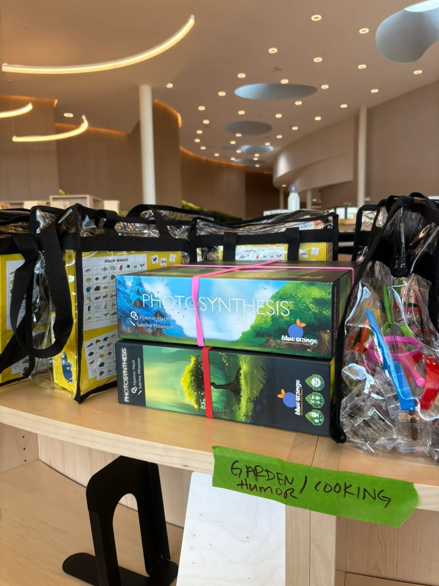 Products displayed on wooden counter in modern interior space, including boxes labeled "Photo Synthesis" and "Blue Orange," black reusable bags, and a green label reading "Garden / Cooking."