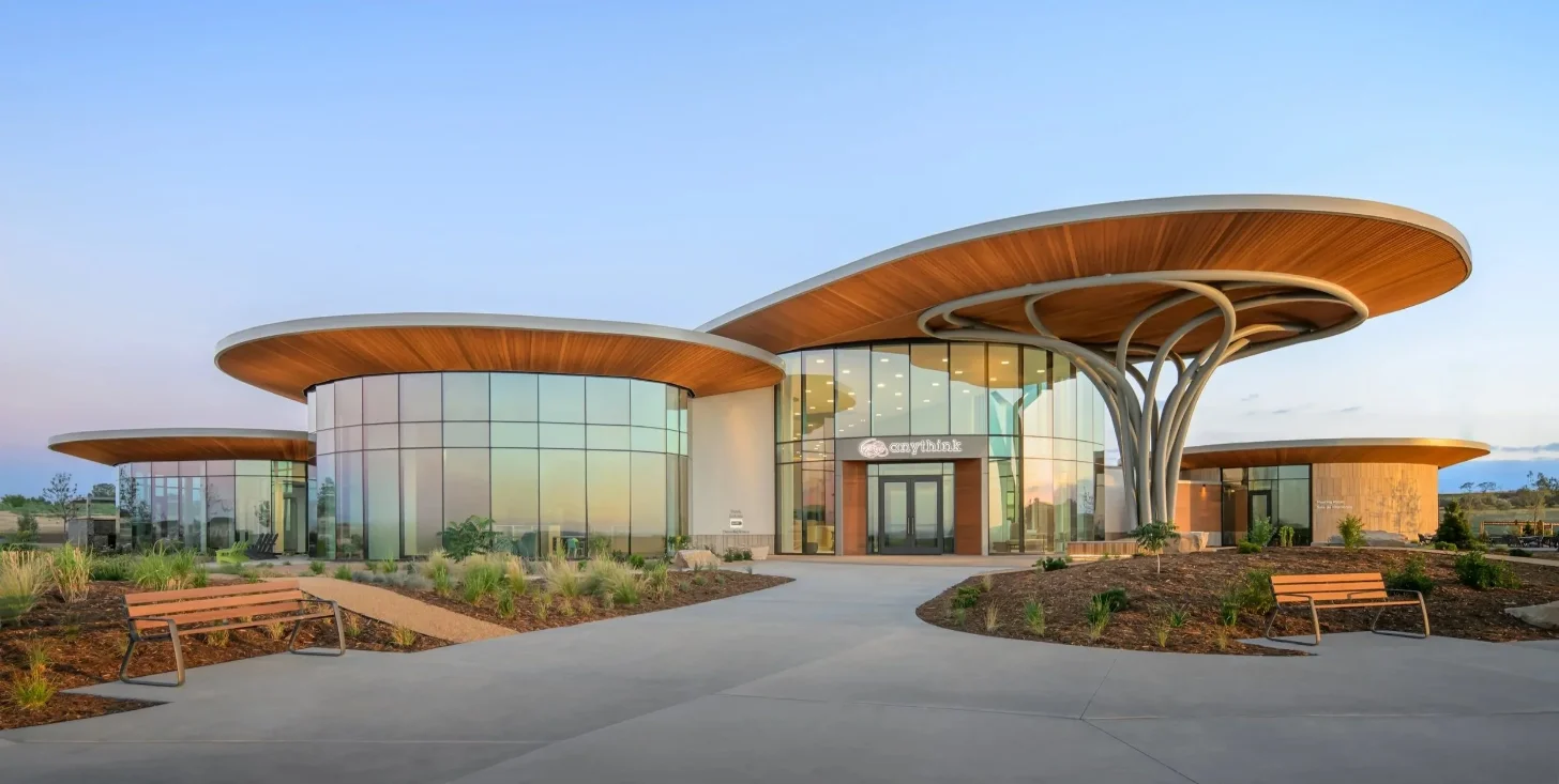 Modern curved building with wooden canopy overhangs and extensive glass walls, flanked by landscaped grounds with native plants and wooden benches along a concrete plaza at dusk.