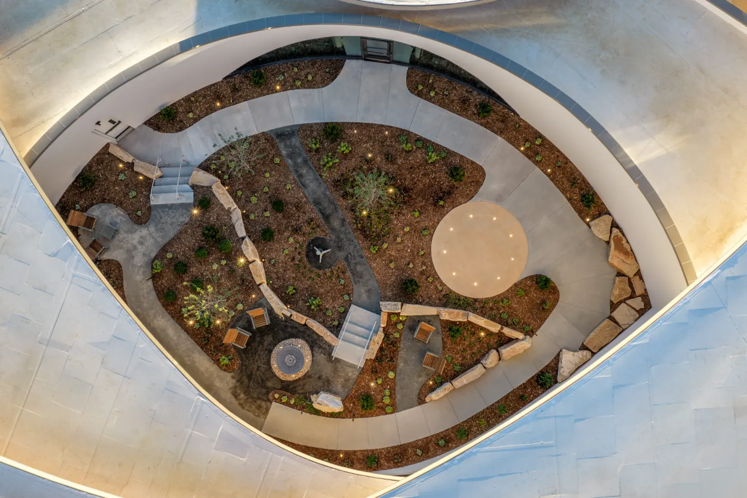 Overhead view of a circular courtyard with concentric rings of landscaping, planted areas, and pathways. Wooden benches and seating areas are arranged around the perimeter, with a large circular table in the center and a circular water feature or plaza element below it. Stone retaining walls and scattered rocks border the outer edge.