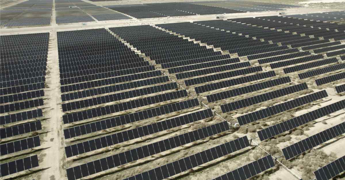 Aerial view of a large solar panel farm with rows of dark photovoltaic panels arranged in parallel lines across flat terrain, separated by light-colored access roads.