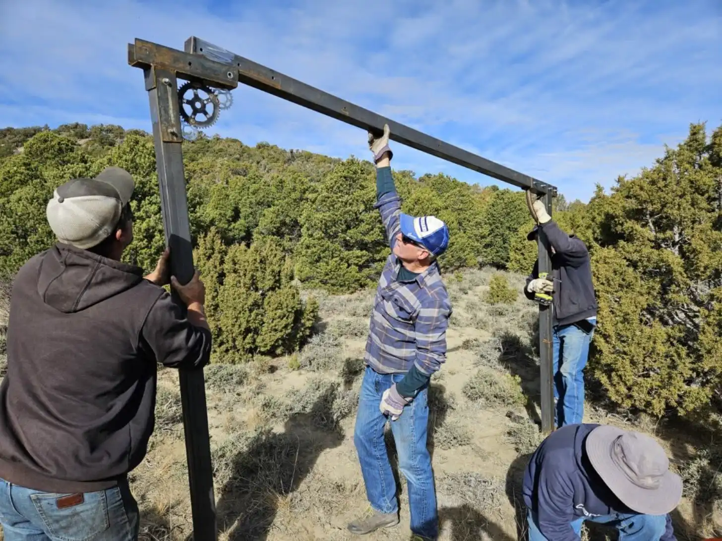 Four people work together to lift a wooden beam or pole structure with a metal gear mechanism attached, standing in a desert landscape with evergreen trees in the background.