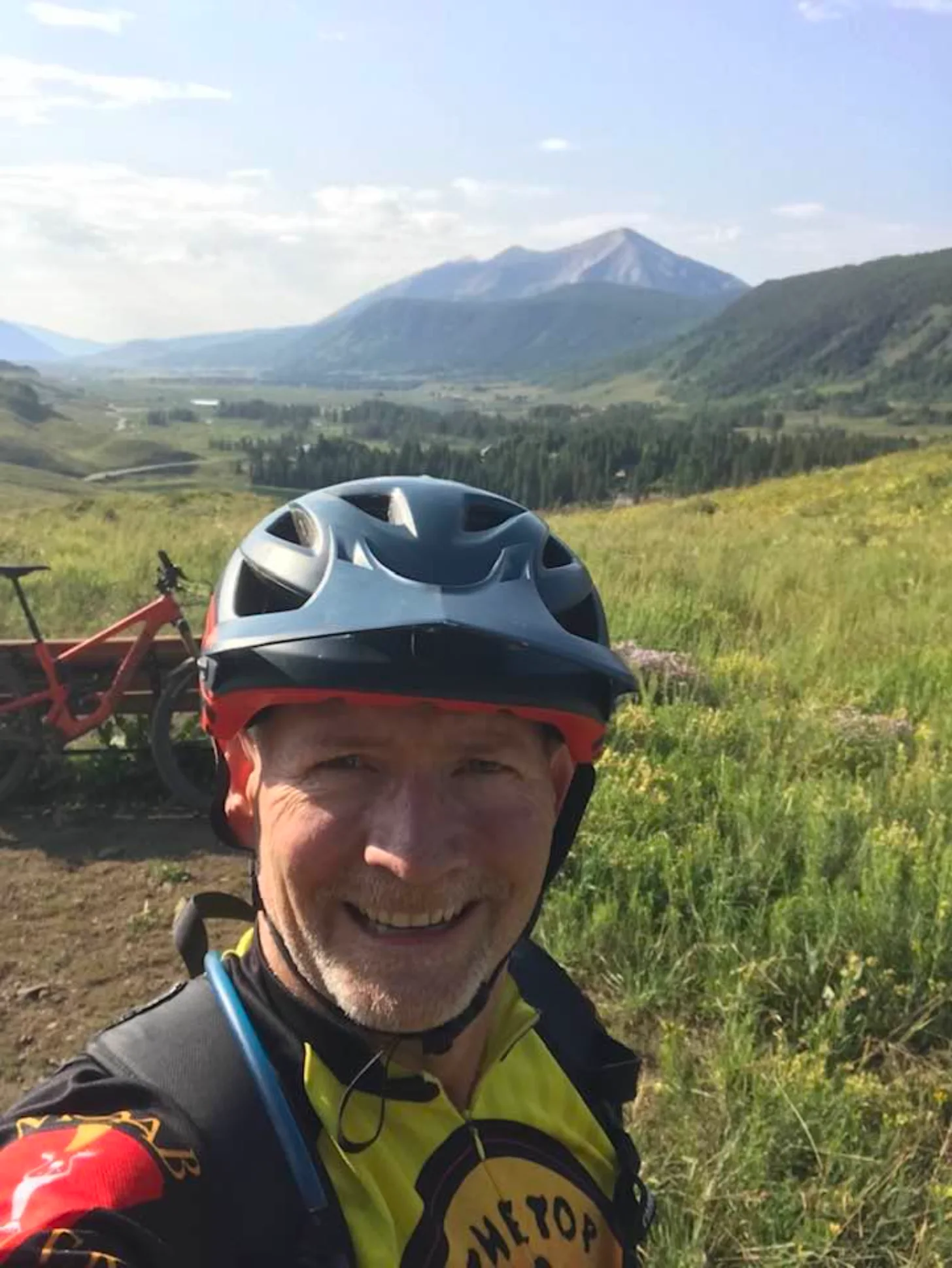 A person wearing a white and black bicycle helmet and yellow safety vest smiles at the camera while standing in a grassy mountain valley. A red mountain bike is visible to the left, with a snow-capped volcano and forested landscape in the background.