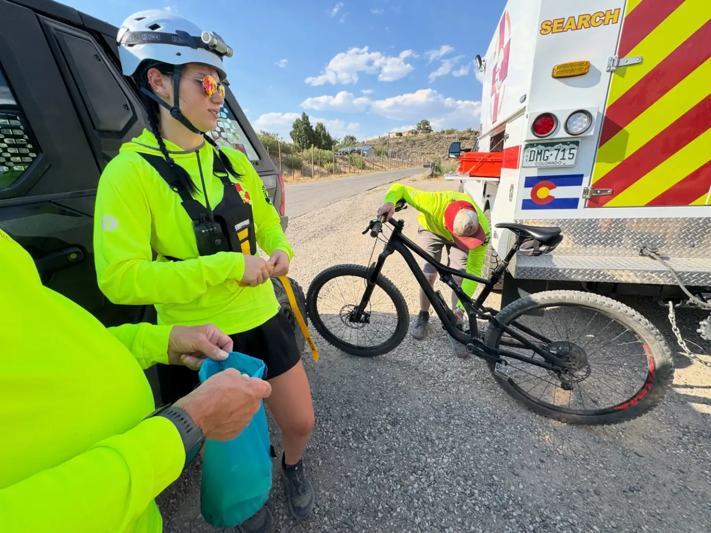 Two emergency responders in high-visibility gear stand beside a black bicycle next to a red and white search and rescue vehicle on a rural road.