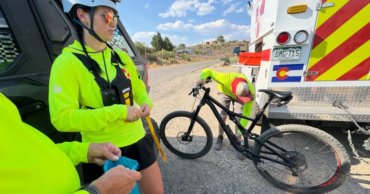 Two people in neon yellow safety vests and helmets stand next to an emergency vehicle with a black bicycle. License plate reads DMG-715. Hillside visible in background.