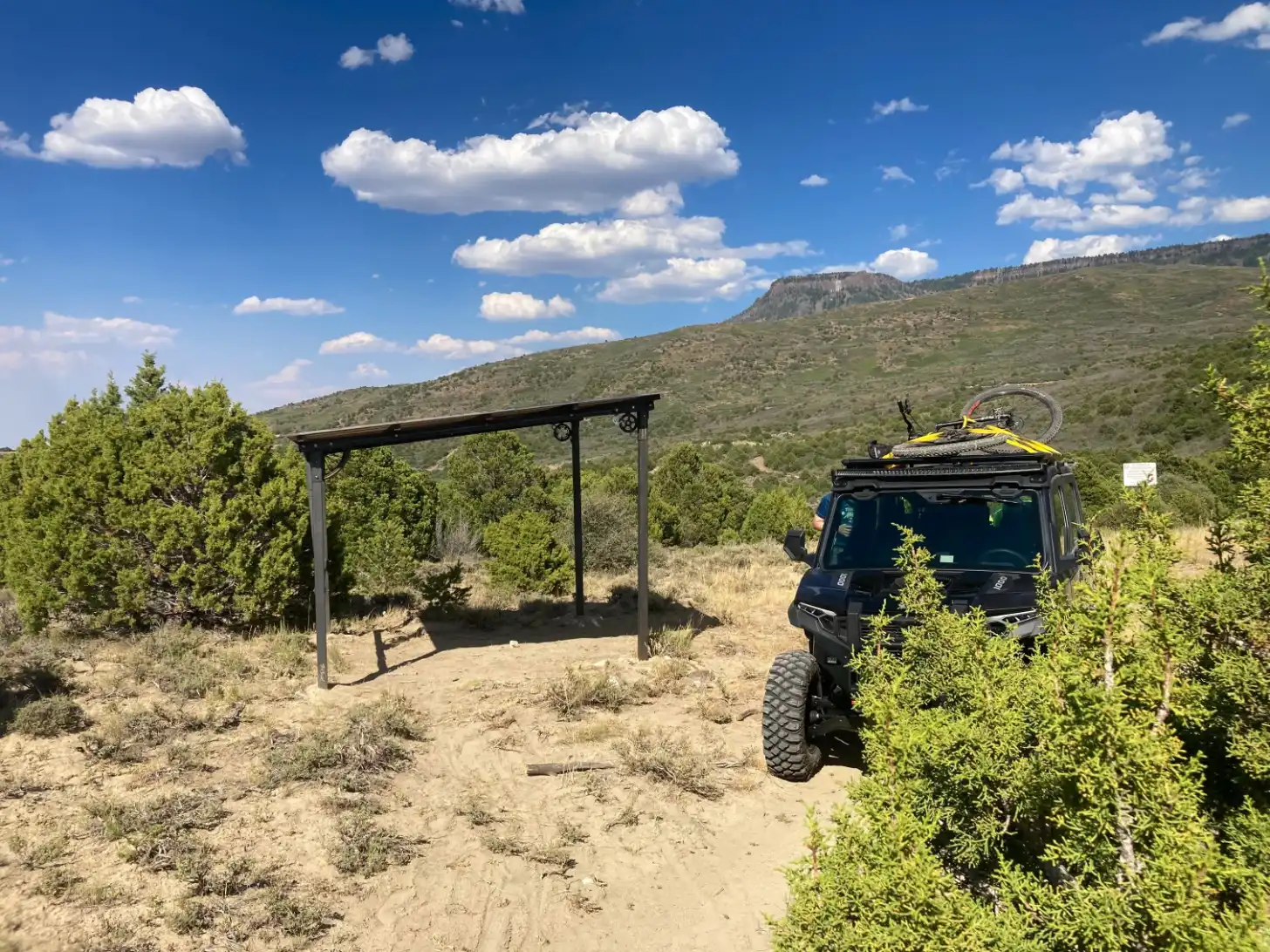A black truck with a yellow mountain bike on its roof rack sits on a dirt road next to a metal-frame shelter structure. Rolling hills covered with scrubland and coniferous trees stretch across the background under a blue sky with white clouds.