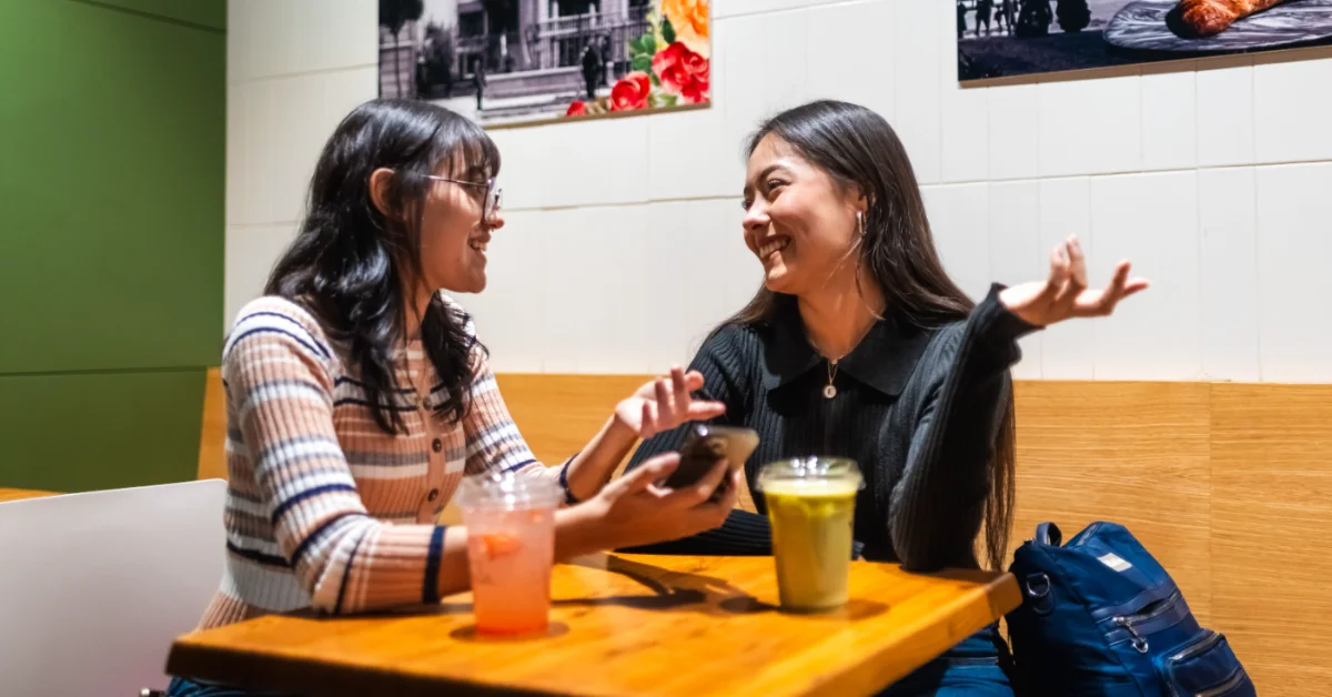 Two women sit at a wooden table facing each other, both smiling and gesturing while talking. One wears a brown and white striped shirt with glasses; the other wears a dark pinstriped shirt. Drinks and a blue bag are on the table. Framed photographs hang on the white wall behind them.