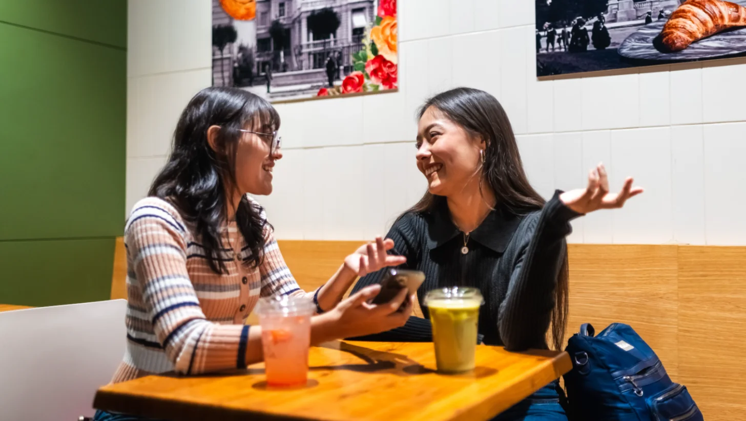 Two women sit at a wooden table facing each other, both smiling and gesturing while talking. One wears a brown and white striped shirt with glasses; the other wears a dark pinstriped shirt. Drinks and a blue bag are on the table. Framed photographs hang on the white wall behind them.