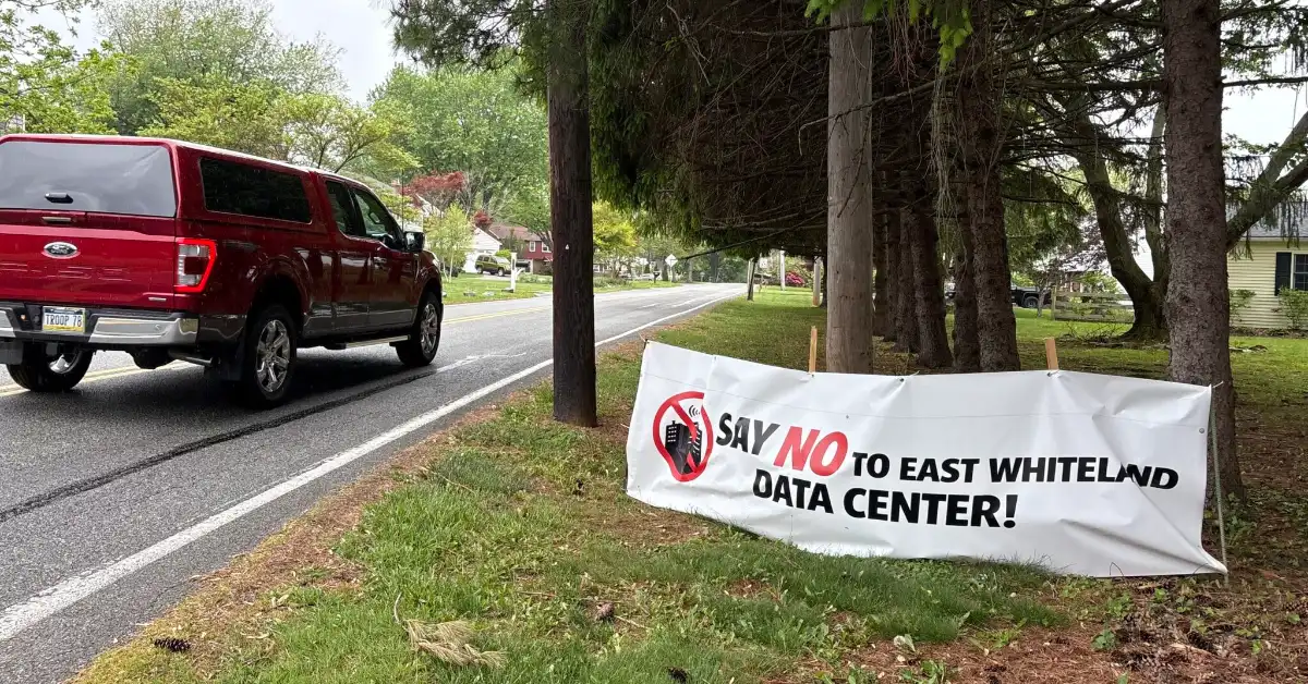 A white banner hanging from trees along a residential street reads "SAY NO TO EAST WHITELAND DATA CENTER!" with a red prohibition symbol. A red pickup truck is parked on the road nearby.