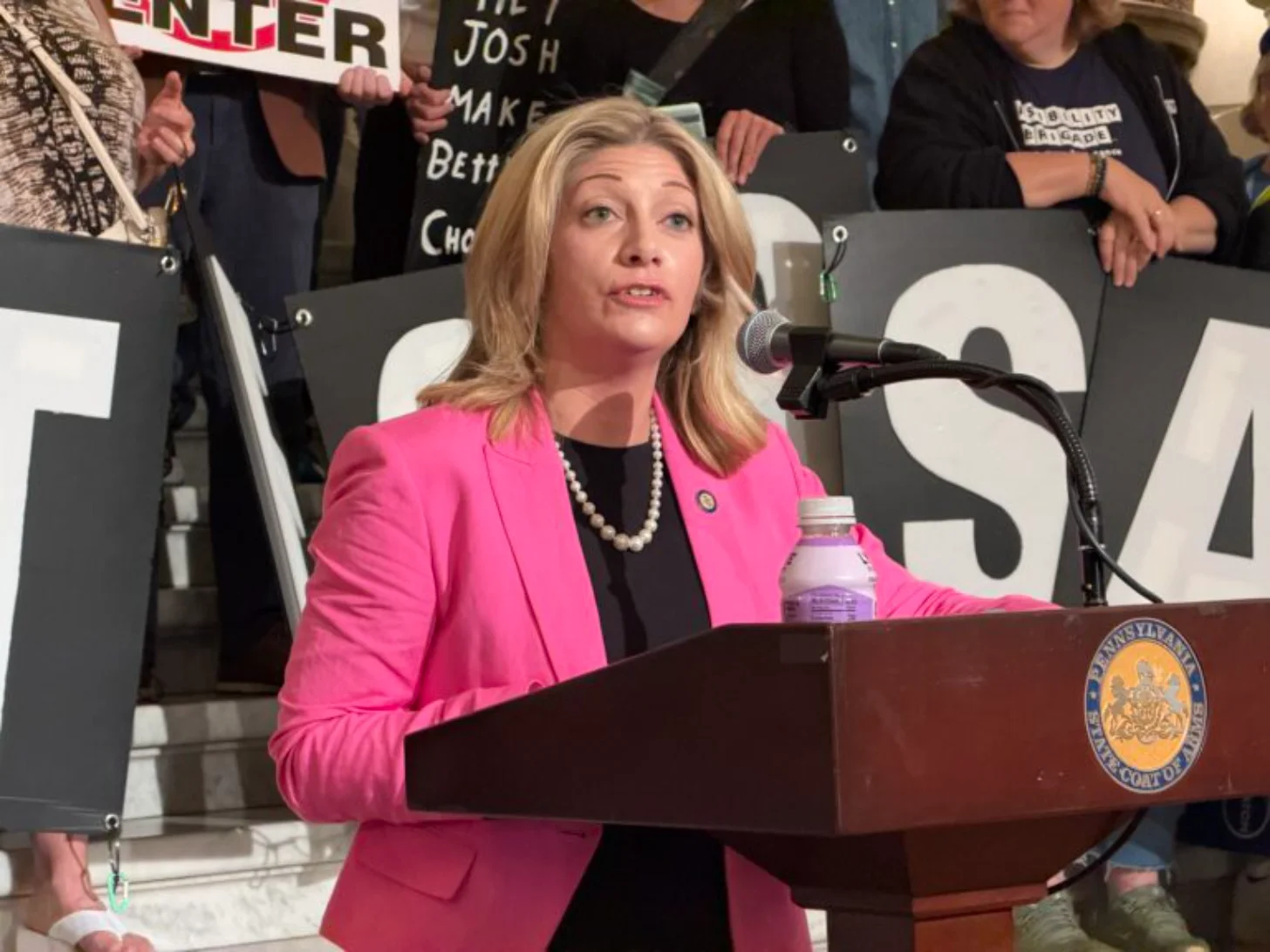 Woman in bright pink blazer speaks at podium with Pennsylvania state seal, standing before supporters holding signs behind her.