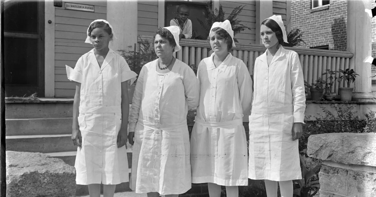 Four Black women in white nursing uniforms and caps stand outside a brick building with a porch, looking toward the camera.