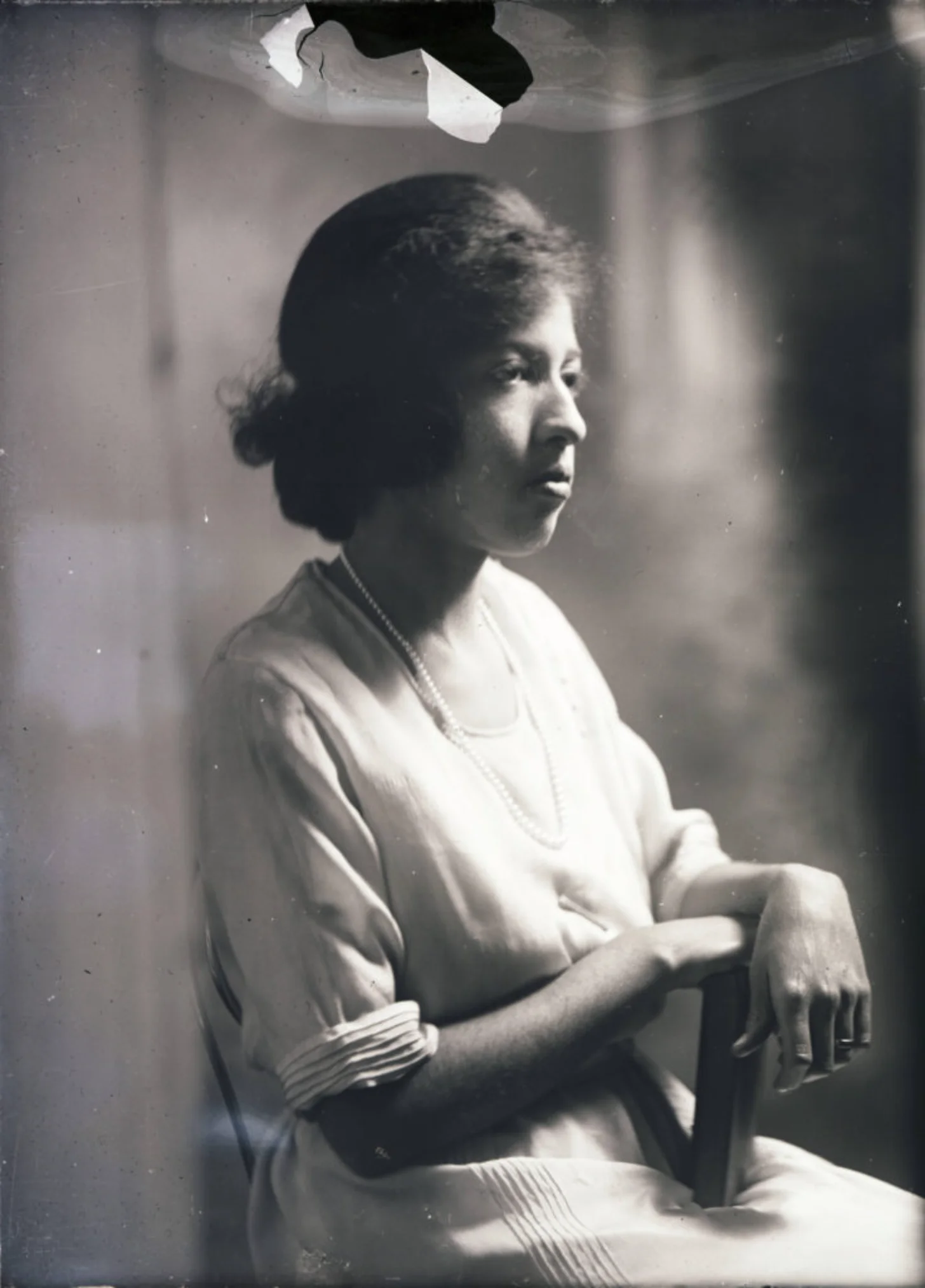 Black and white portrait photograph of a Black woman in profile, seated and wearing a light-colored dress with pleated sleeves and a beaded necklace.