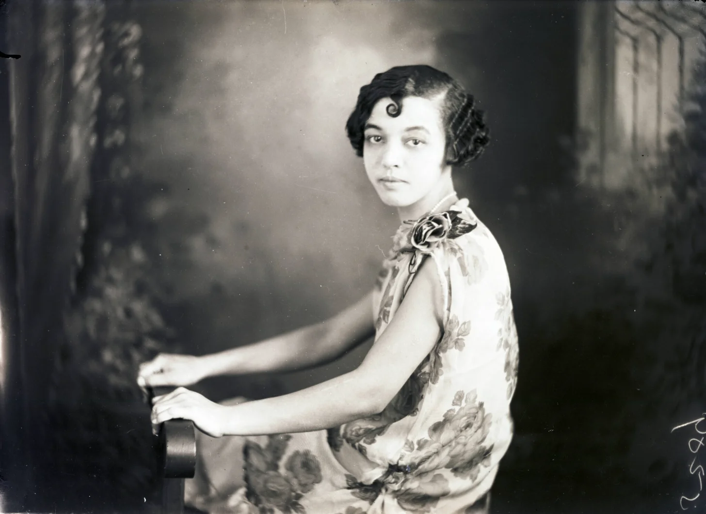 A Black woman in a light-colored dress with floral embroidery sits at a piano, looking toward the camera over her shoulder. Her dark hair is styled in waves with a decorative curl on her forehead.