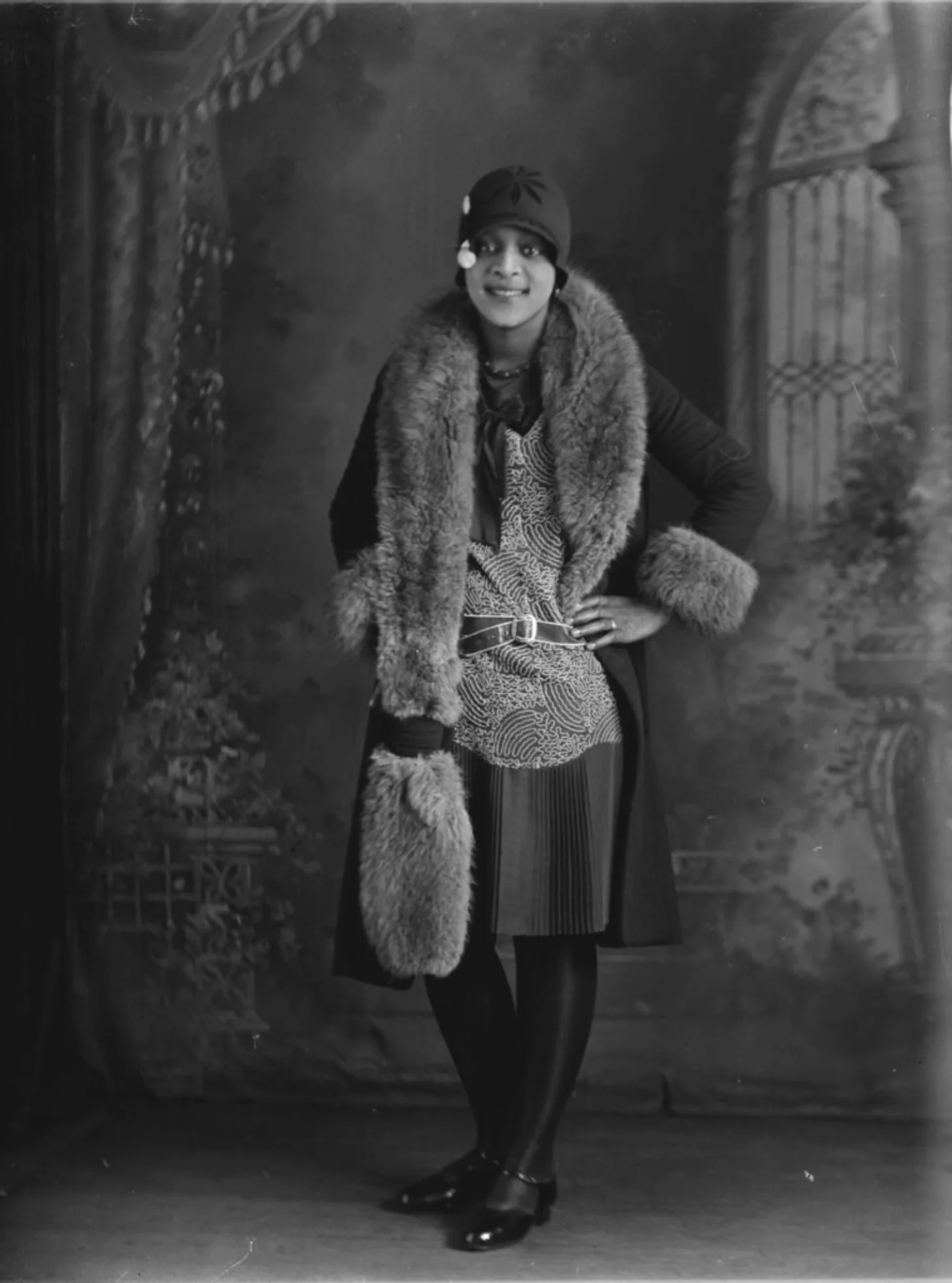 A Black woman in 1920s-style dress with fur stole and cloche hat stands in an ornate interior, hand on hip, smiling at camera.