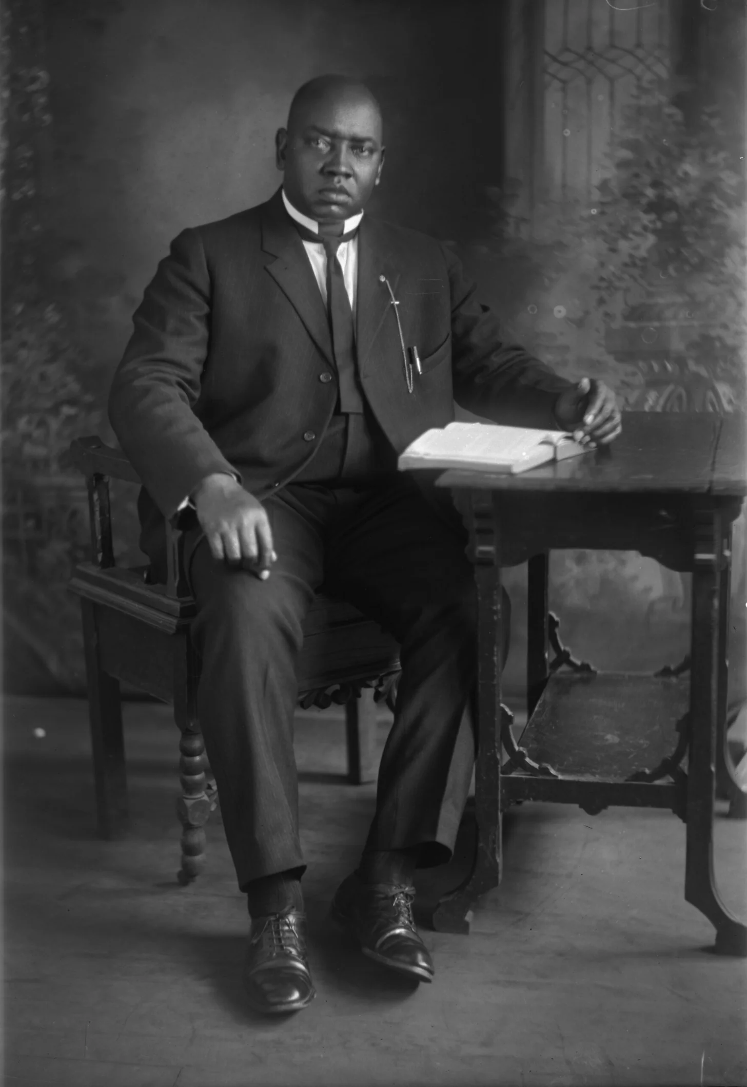 A Black man in a dark suit and white shirt sits at a wooden table holding an open book or document, looking toward the camera in this black and white photograph.