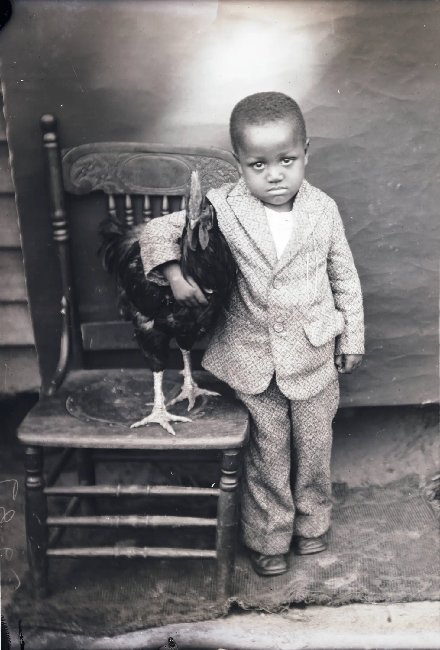 A young child in a light-colored suit stands beside a wooden chair holding a black bird with visible yellow feet.
