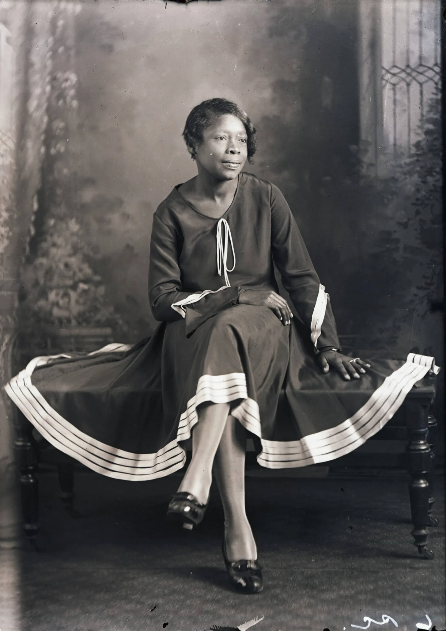 A Black woman seated on a chair wears a dark dress with white trim at the neckline and cuffs, and white-trimmed ruffles on the skirt. She gazes to the side while resting one hand on her lap.