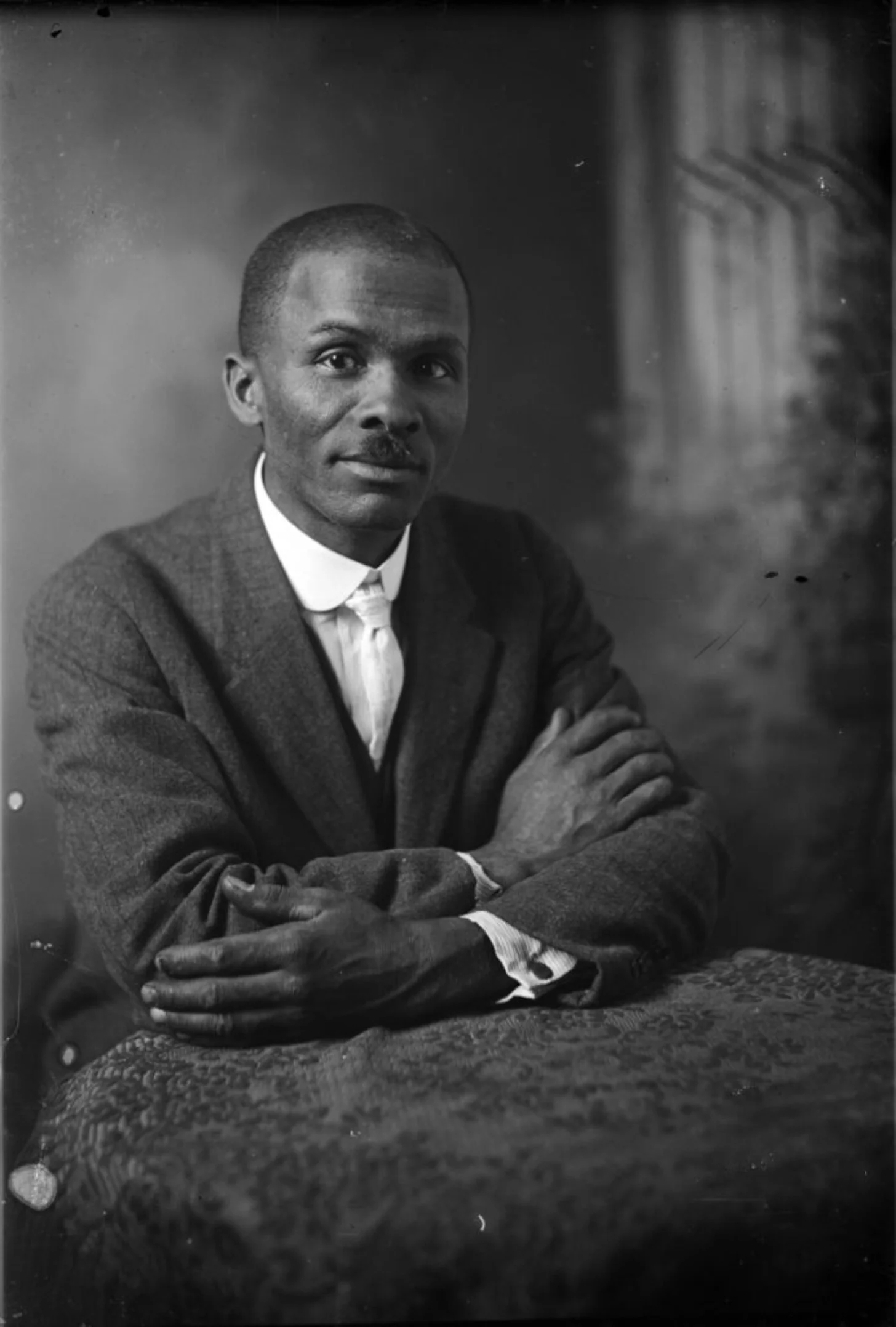 Black and white portrait photograph of a Black man in formal attire with a dark suit jacket and white shirt, seated with his arms crossed, looking toward the camera.