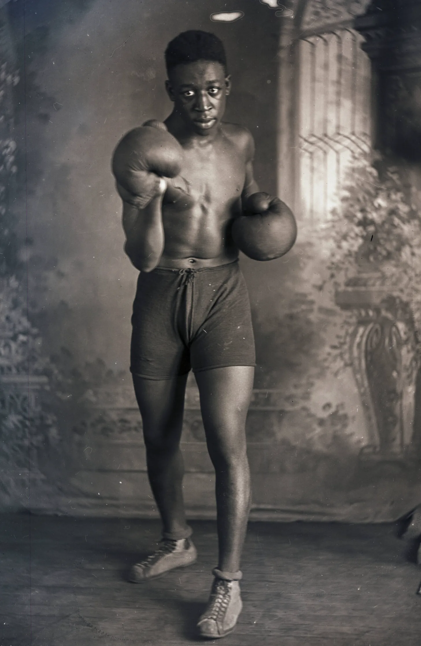 A Black man, shirtless and wearing boxing gloves and shorts, stands in fighting stance in an indoor studio setting with draped fabric backdrop.