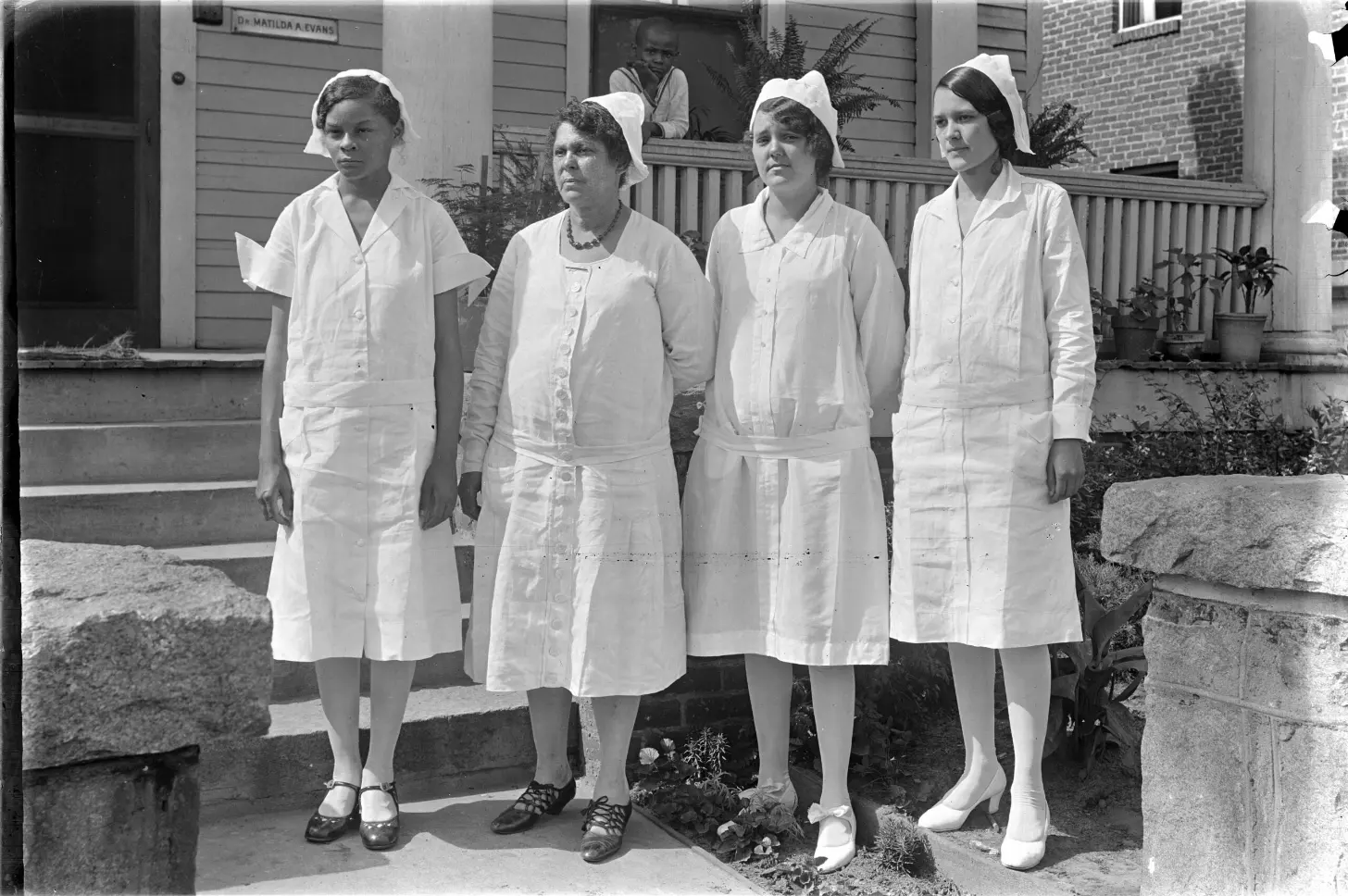 Four Black women in white uniforms and white head coverings stand outside a brick building with a sign reading "Dr. Matilda A. Evans" visible above the entrance.