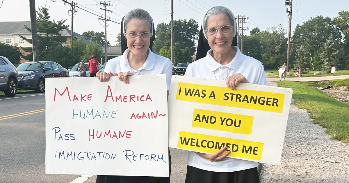 Two nuns hold signs on a residential street. The left sign reads "Make America HUMANE again~ Pass HUMANE IMMIGRATION REFORM" in red and blue handwriting. The right sign has yellow highlighted text reading "I WAS A STRANGER AND YOU WELCOMED ME."