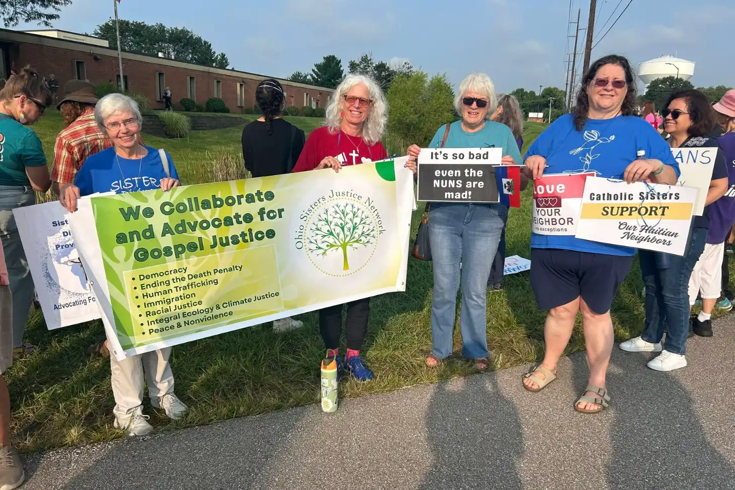 A group of people stand outdoors holding signs and banners, including one that reads "We Collaborate and Advocate for Gospel Justice" with a tree logo, another stating "It's so bad even the NUNS are mad," and a third reading "Catholic Sisters SUPPORT our Haitian Neighbors." A brick building and grassy field are visible in the background.