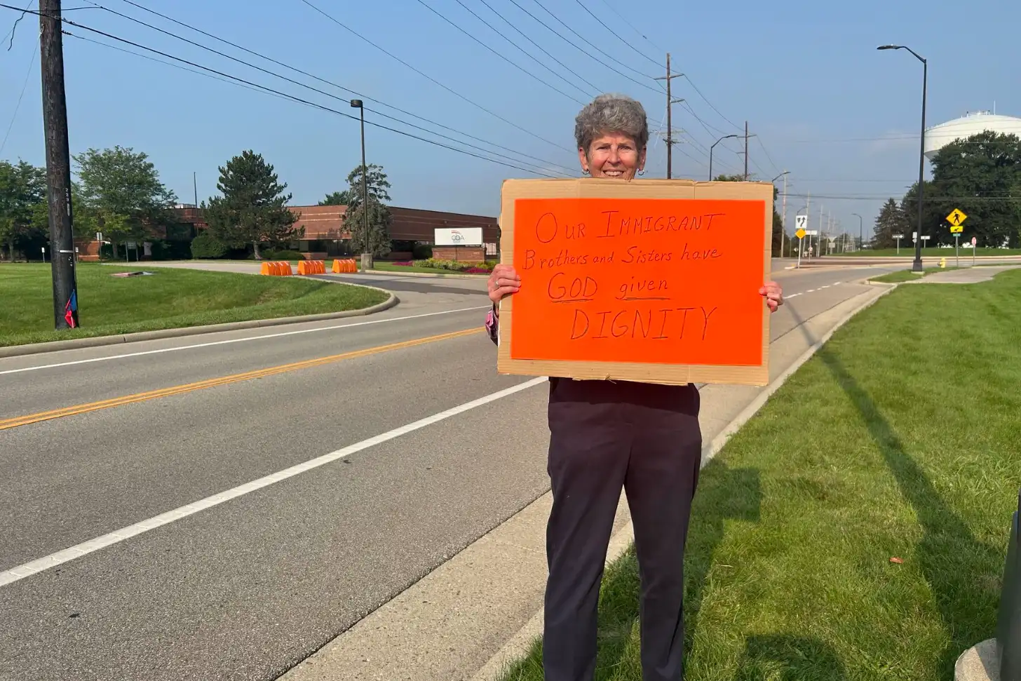 A woman holds an orange sign reading "Our Immigrant Brothers and Sisters have GOD given DIGNITY" while standing on a sidewalk along a road with buildings and power lines visible in the background.