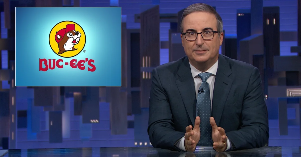 Man in dark suit and glasses, John Oliver, speaks at desk with Buc-ee's logo displayed on screen behind him against a cityscape backdrop.