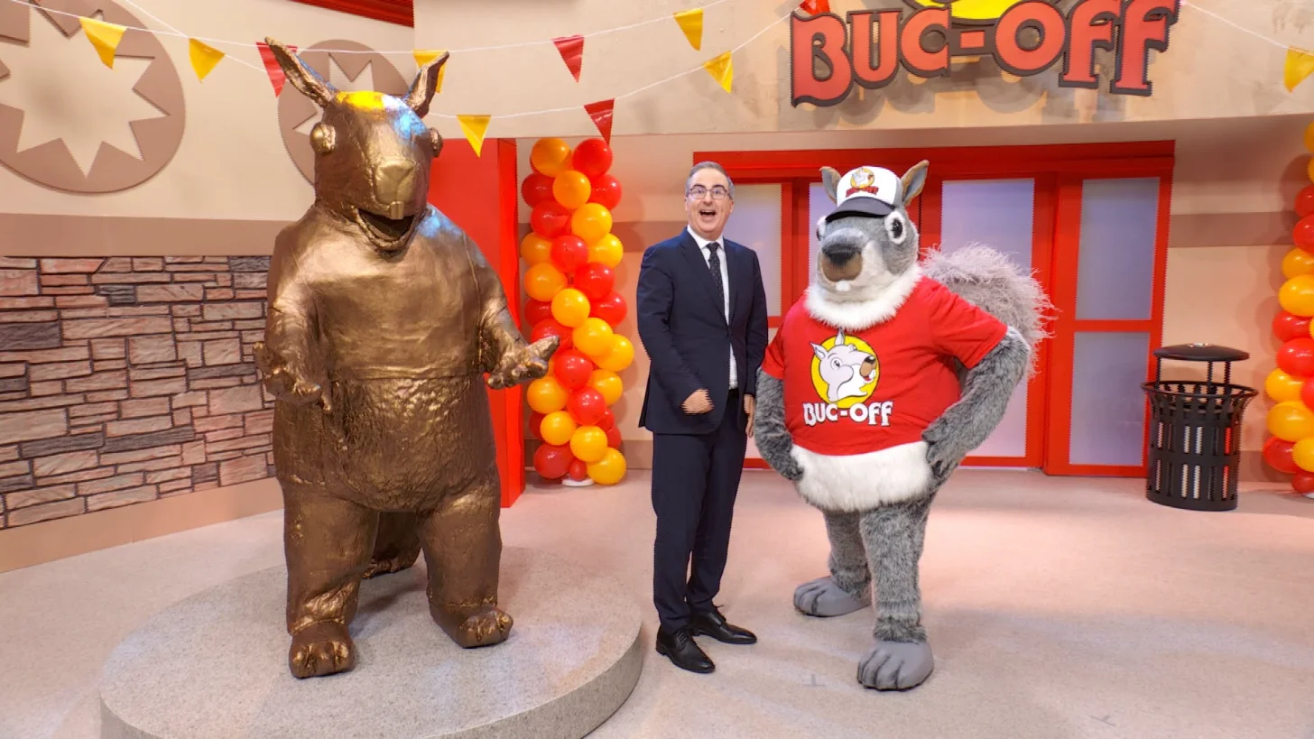 A man in a dark suit, John Oliver, stands between a large bronze squirrel statue and a squirrel mascot character in a red shirt labeled "BUC-OFF" on a studio set decorated with red and yellow balloons and bunting.