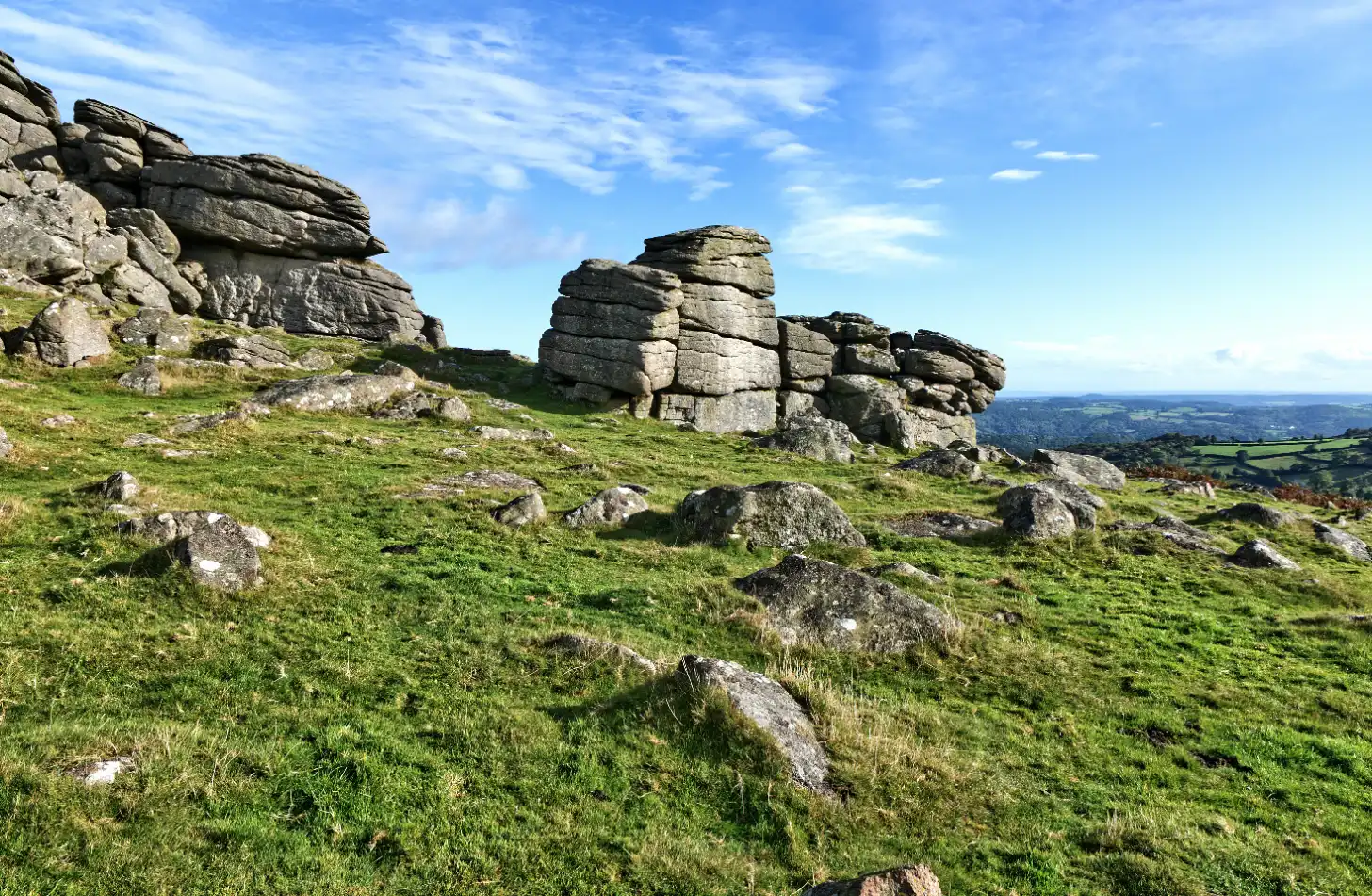 Stacked granite rock formations rise from a grassy hillside with scattered boulders, overlooking a patchwork of fields and woodland valleys below under a partly cloudy sky.