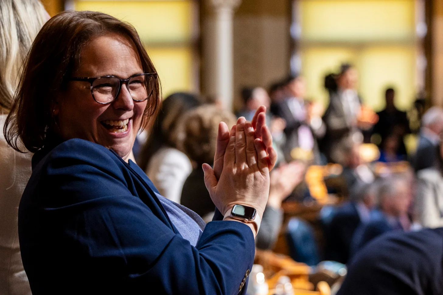 Woman wearing glasses and a dark blue blazer claps her hands while smiling at a crowded indoor event with yellow-lit windows in the background.