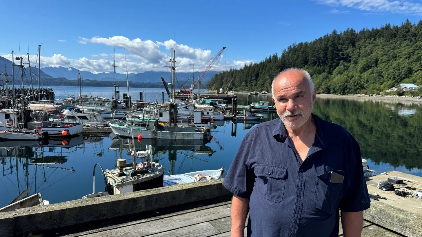 A man in a dark blue work shirt stands on a wooden dock facing the camera. Behind him is a harbor filled with fishing boats moored at wooden pilings, with forested hills and snow-capped mountains visible across the water under a partly cloudy sky.