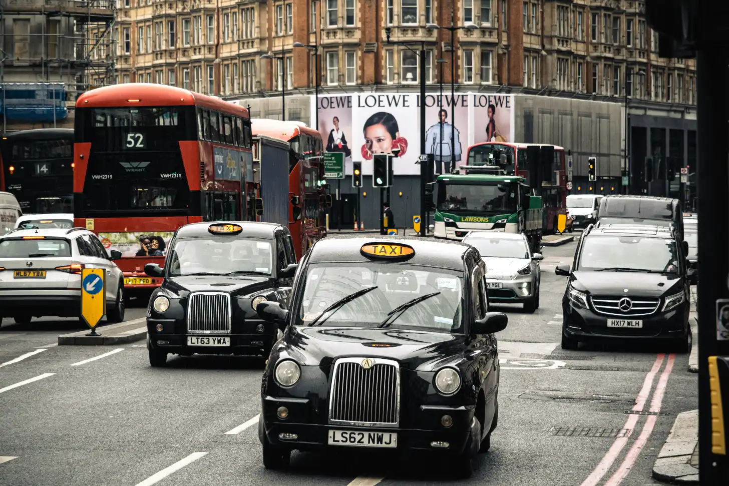 Black taxi cabs and a red double-decker bus on a London street, with Victorian buildings and LOEWE fashion advertisements in the background.