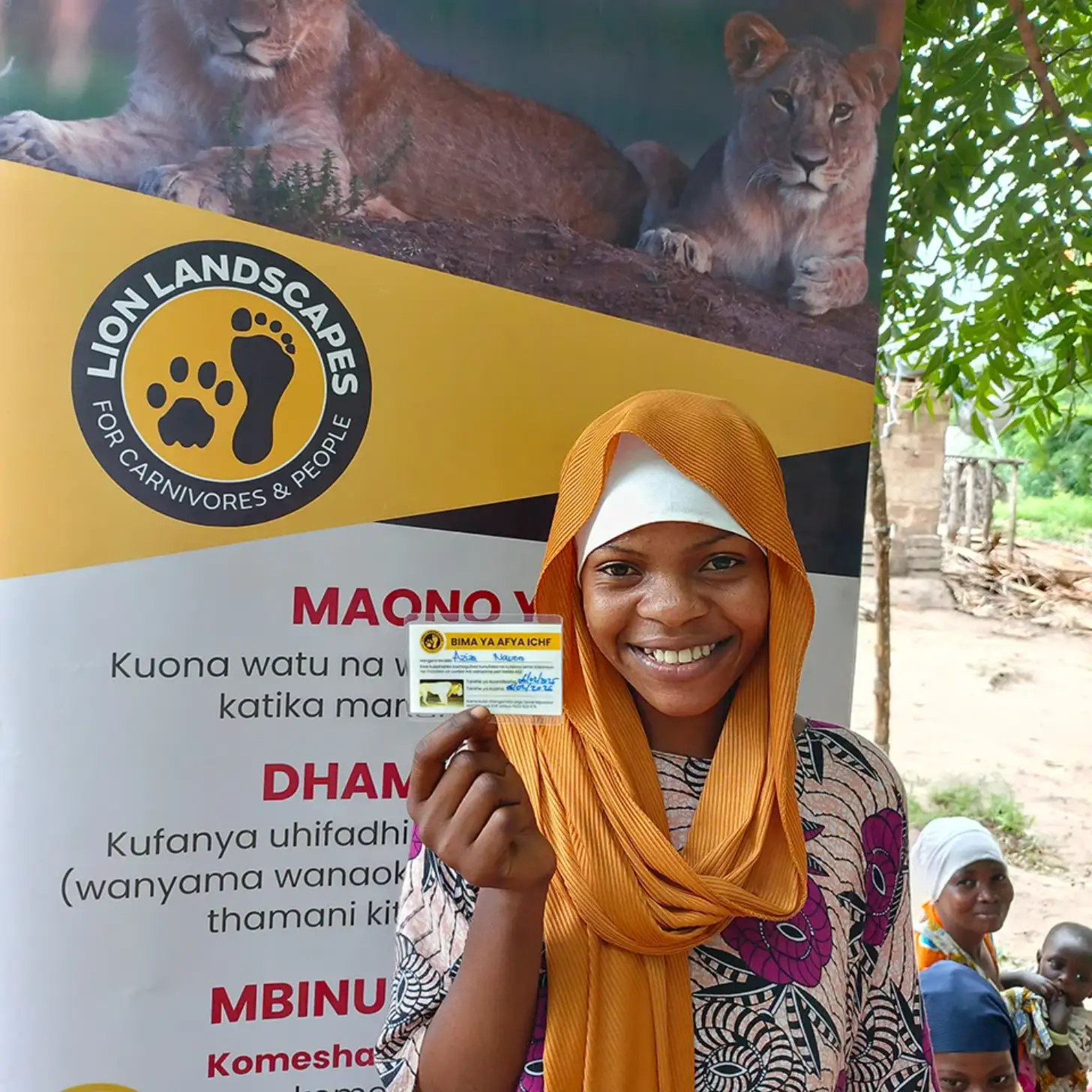 A young woman wearing an orange headscarf smiles while holding up a card in front of a banner that reads "LION LANDSCAPES FOR CARNIVORES & PEOPLE" with images of lions at the top.