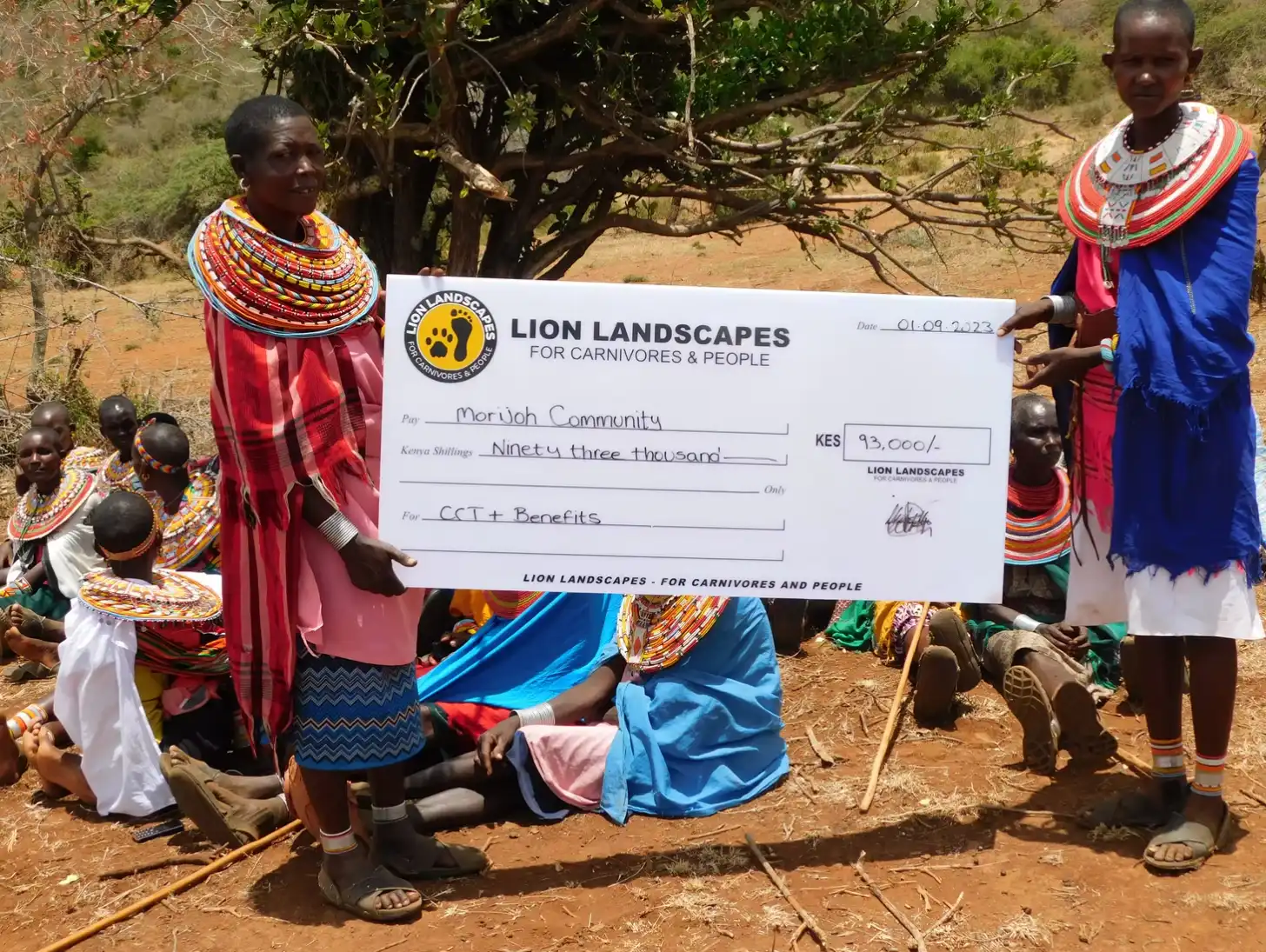 Members of the Morudi Community hold a check from Lion Landscapes for 93,000 Kenya Shillings dated 01.09.2023, designated for CCT+ Benefits, in an outdoor setting with trees and dry ground.