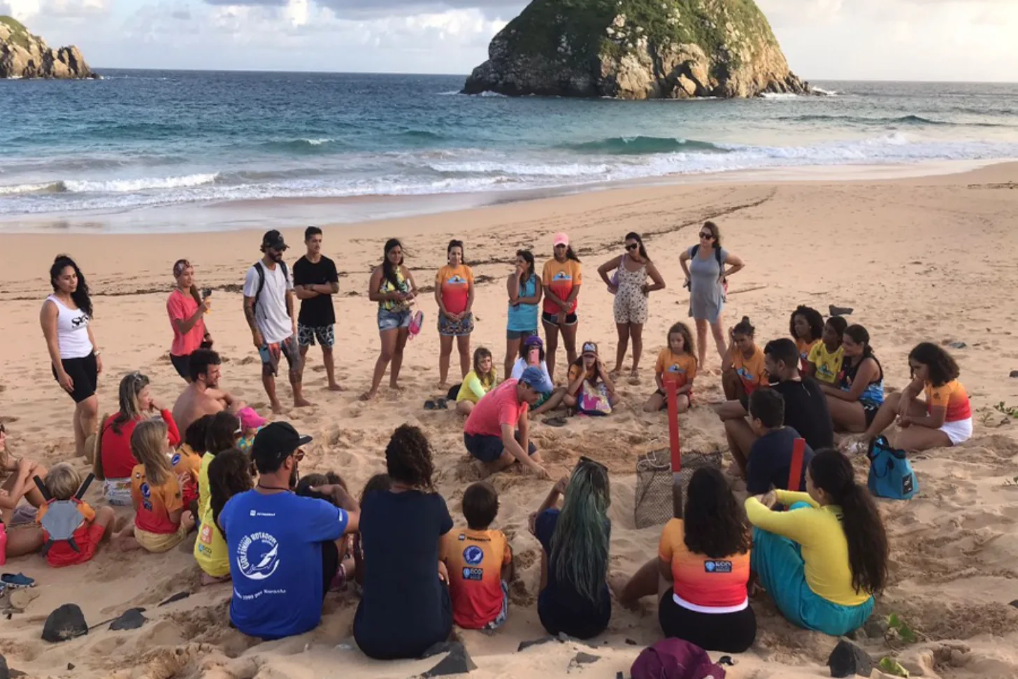 A large group of people gathered in a circle on a sandy beach, with a large rocky island visible in the ocean behind them.