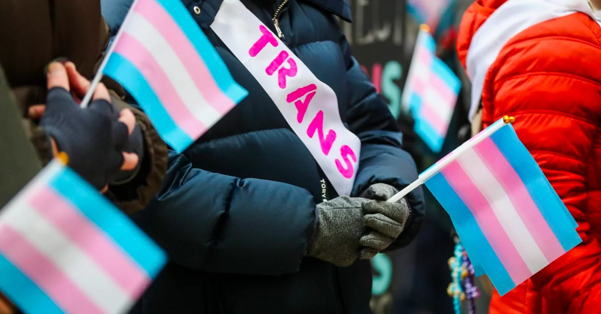 People wearing winter coats hold trans pride flags and a sign reading "TRANS" at an outdoor gathering.