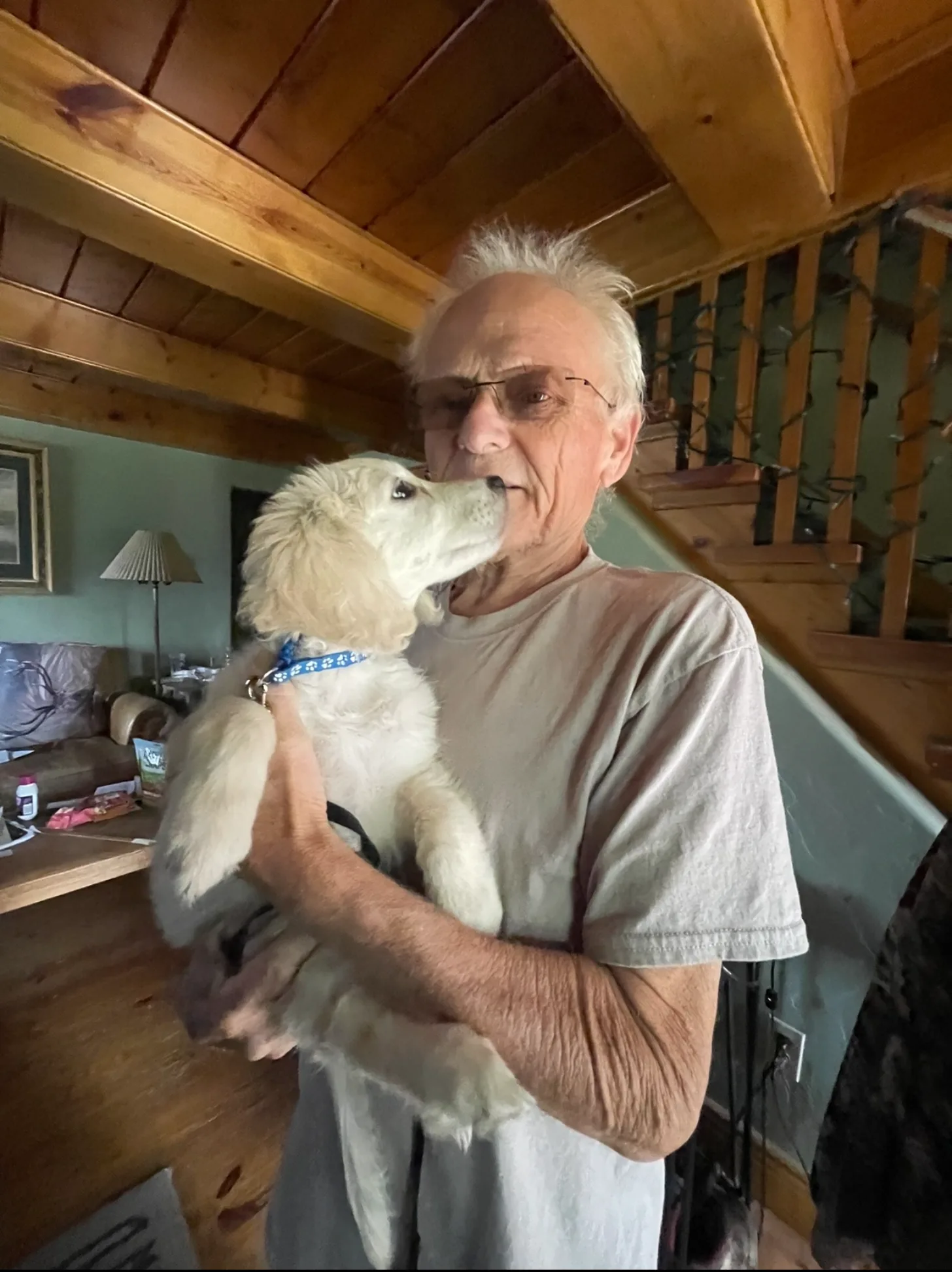 An older man wearing glasses and a tan shirt holds a white dog with a blue collar inside a wooden-beamed cabin. The dog's head is raised toward the man's face.