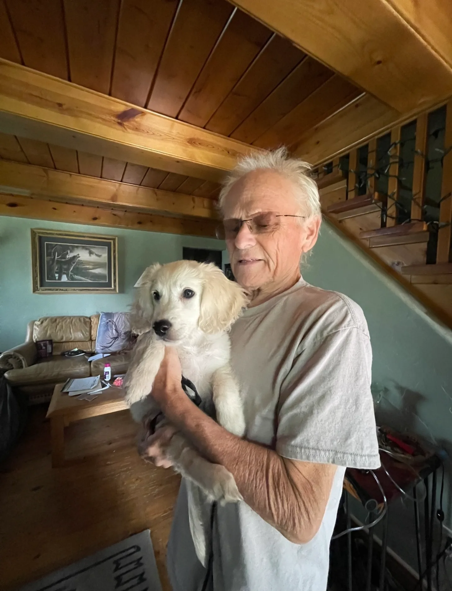 An older man wearing glasses and a tan shirt holds a white dog with long ears in a wooden-beamed room with light blue walls and wooden stairs visible in the background.