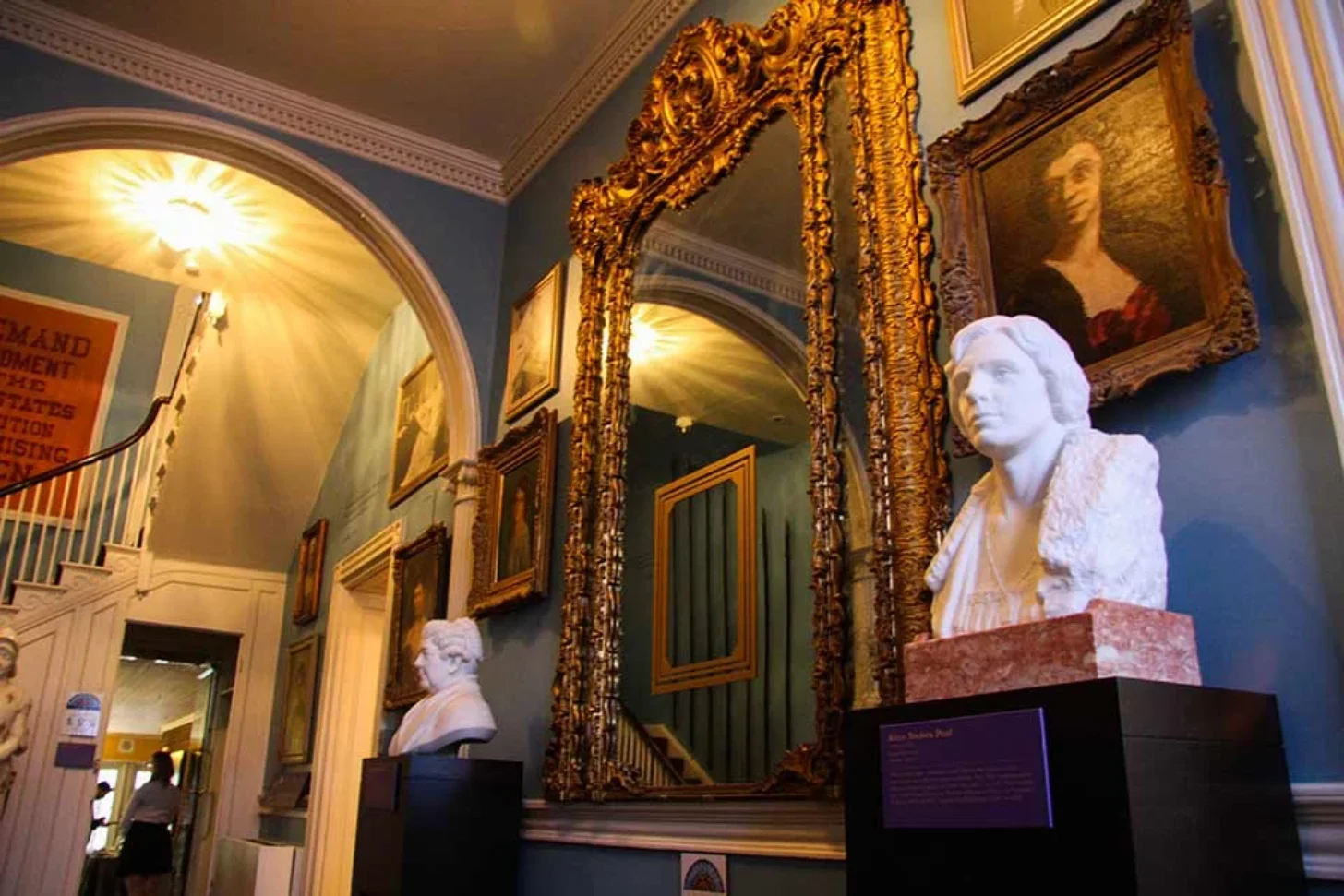 Two white marble busts displayed on pedestals in an ornate gallery hall with gold-framed mirrors and paintings on blue walls.