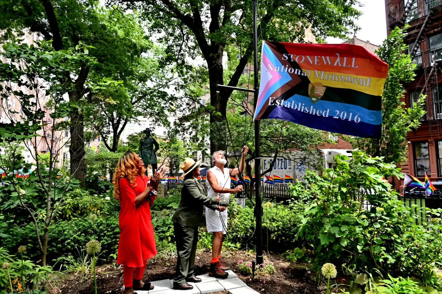 Three people hold a flag reading "STONEWALL National Monument Established 2016" in a garden surrounded by trees and brick buildings.