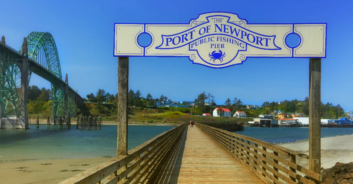 A wooden pier extends into blue-green water beneath a sign reading "THE PORT OF NEWPORT PUBLIC FISHING PIER" with a crab illustration. A steel bridge spans the water to the left, with forested hills and residential buildings visible across the water.