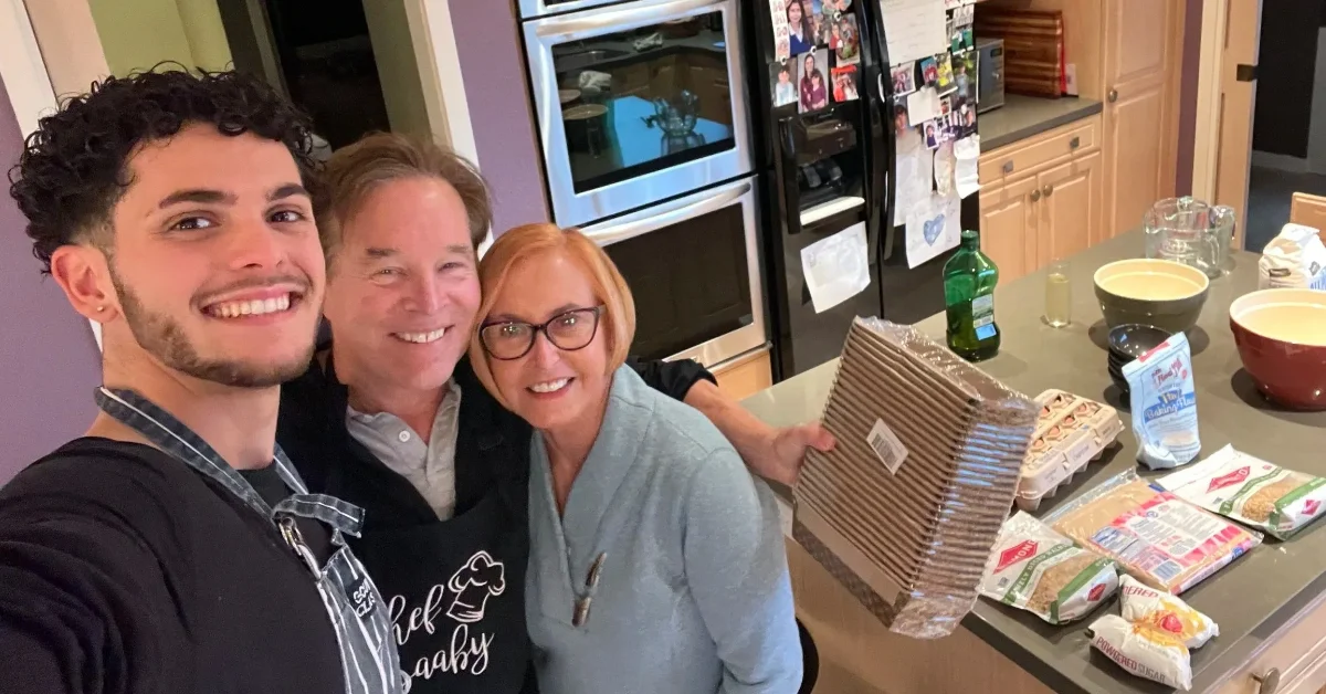 Three people smile at a kitchen counter with packaged foods, bottles, and bowls visible.