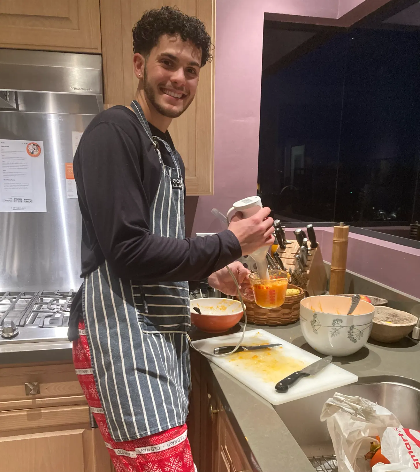 A man in a striped apron stands at a kitchen counter holding a bowl, smiling at the camera. Various cooking ingredients and bowls are arranged on the counter in front of him.