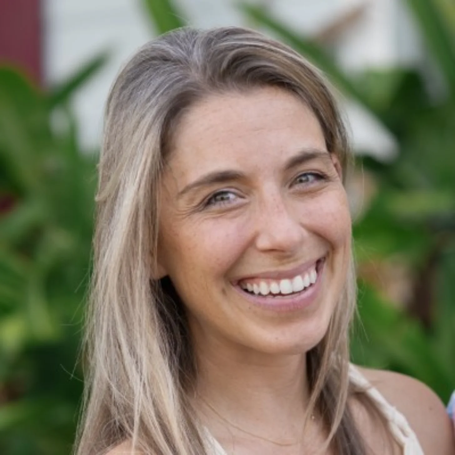 Woman with shoulder-length blond hair smiling at the camera outdoors with green foliage in the background.