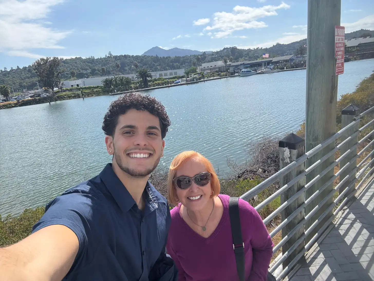 Two people smile at the camera on a wooden viewing platform overlooking a bay with industrial buildings, forested hills, and a mountain in the background under partly cloudy sky.