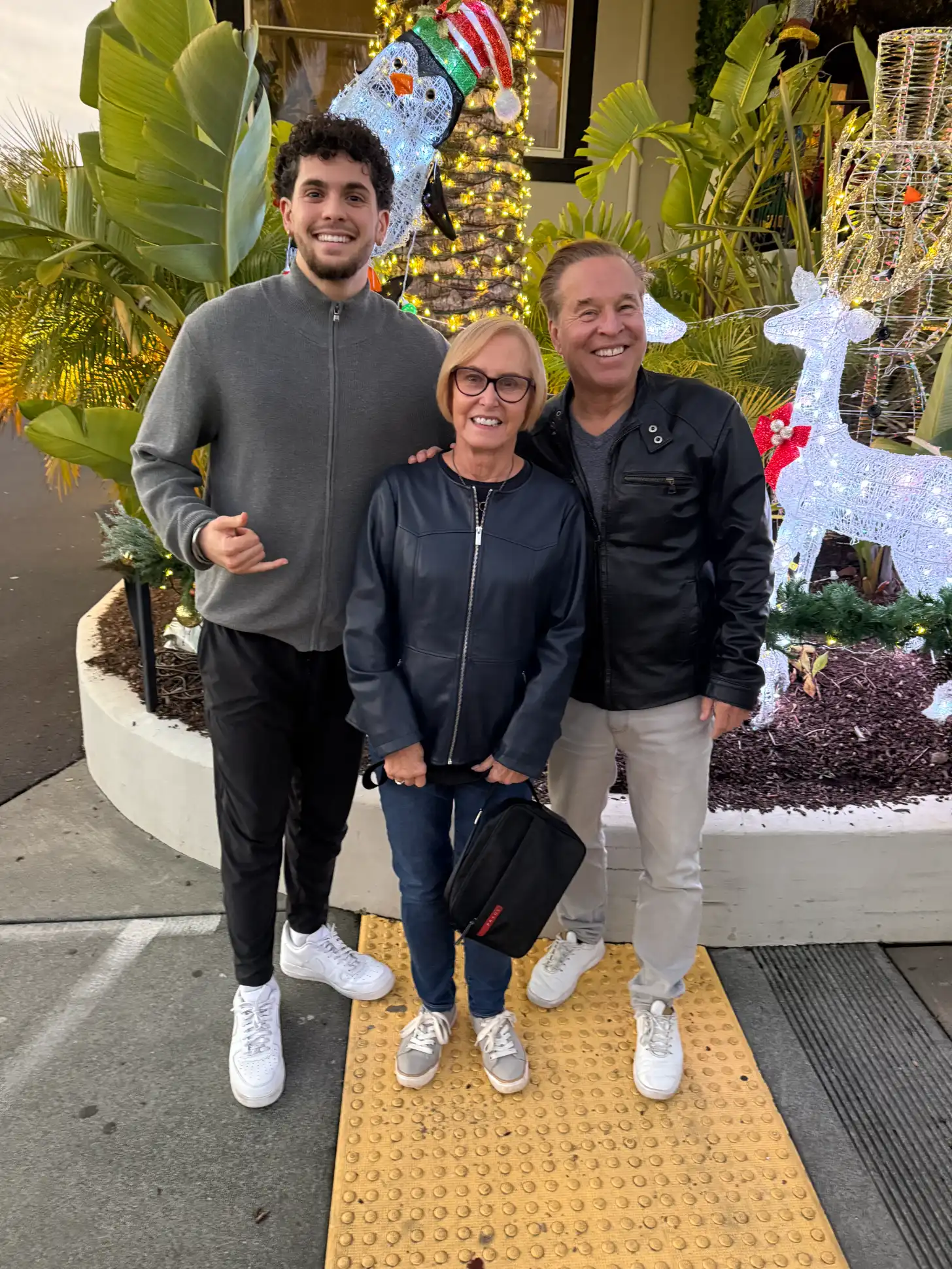 Three people stand smiling on a yellow tactile paving surface in front of a storefront decorated with illuminated holiday displays, including a penguin figure wearing a Santa hat and white reindeer sculptures surrounded by tropical plants.