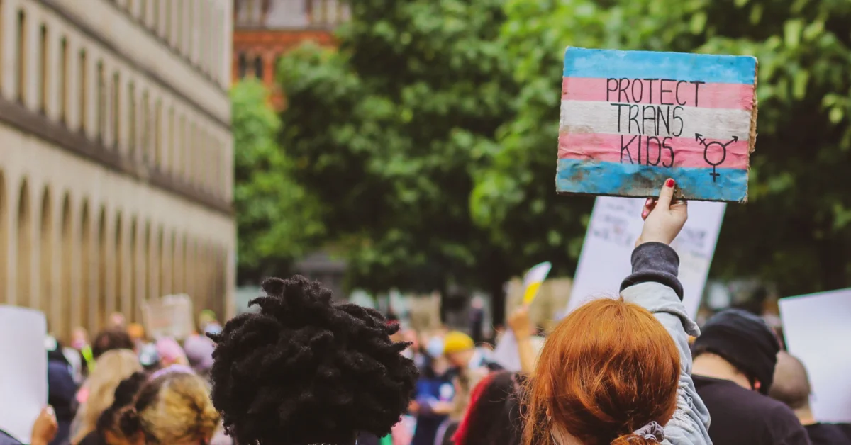 A person with reddish-brown hair holds a hand-painted sign reading "PROTECT TRANS KIDS" in a crowd of protesters on a city street lined with buildings and trees.